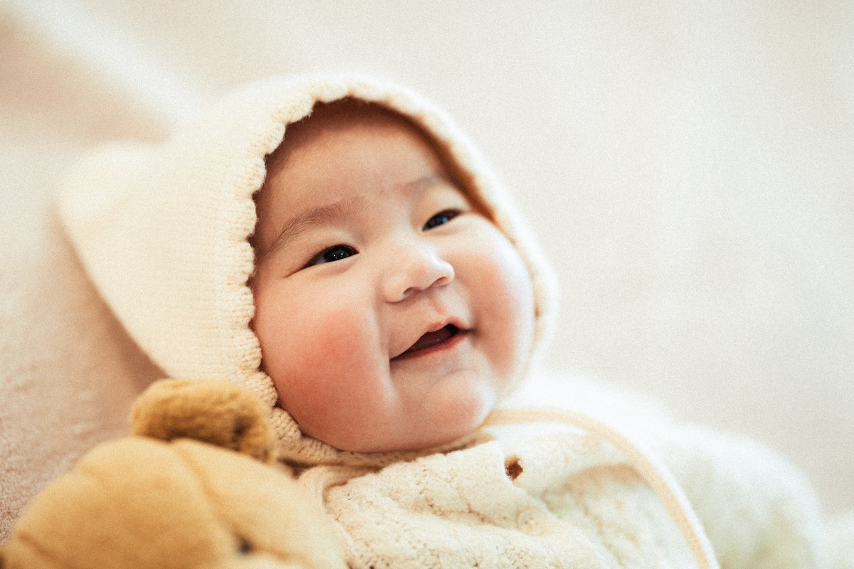 Smiling baby in a cream knit bonnet beside a teddy bear.
