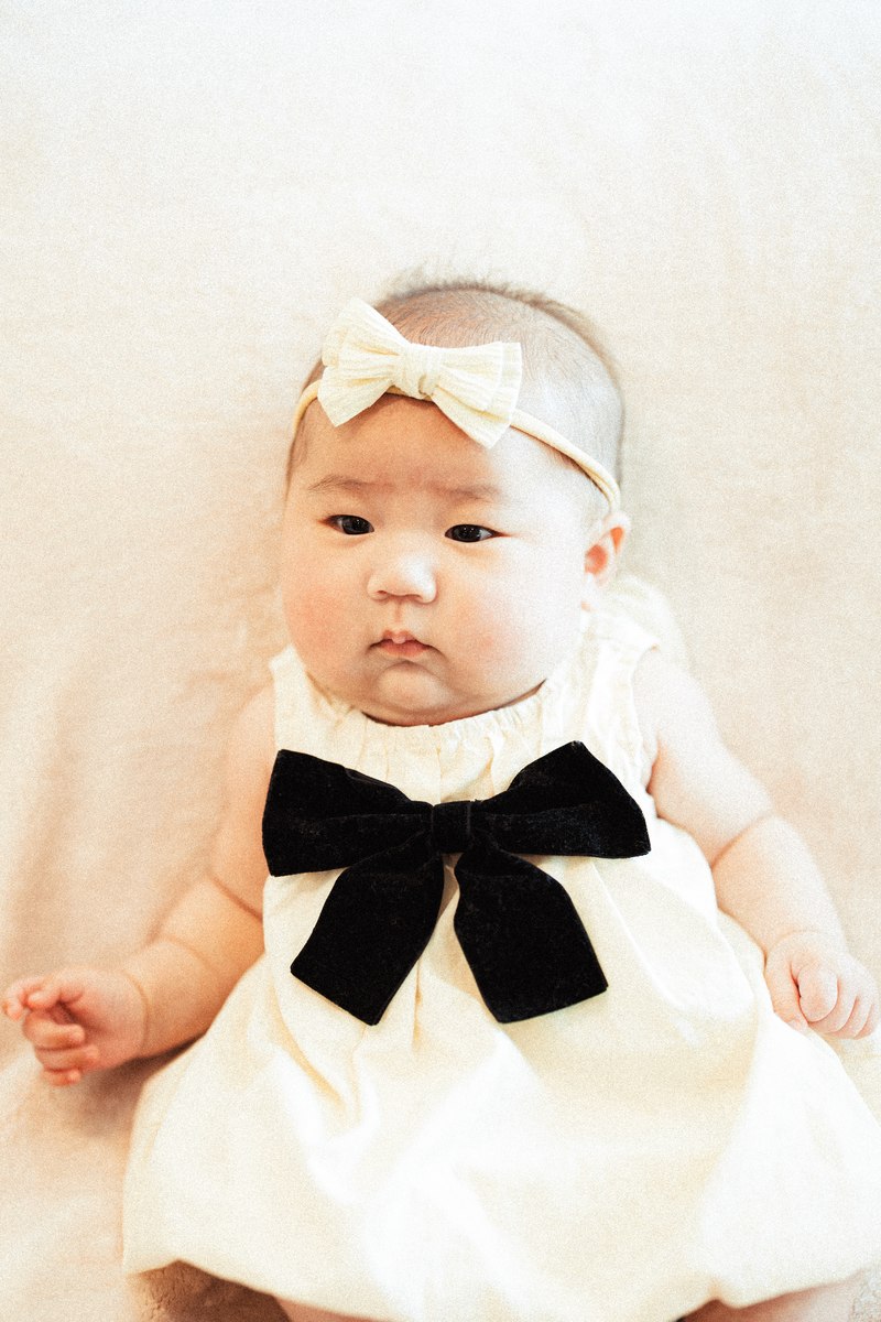 Baby wearing a white dress, large black bow, and cream bow headband.